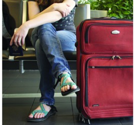 girl wearing jeans and flipflops sitting on an airport seat with a red carryon bag